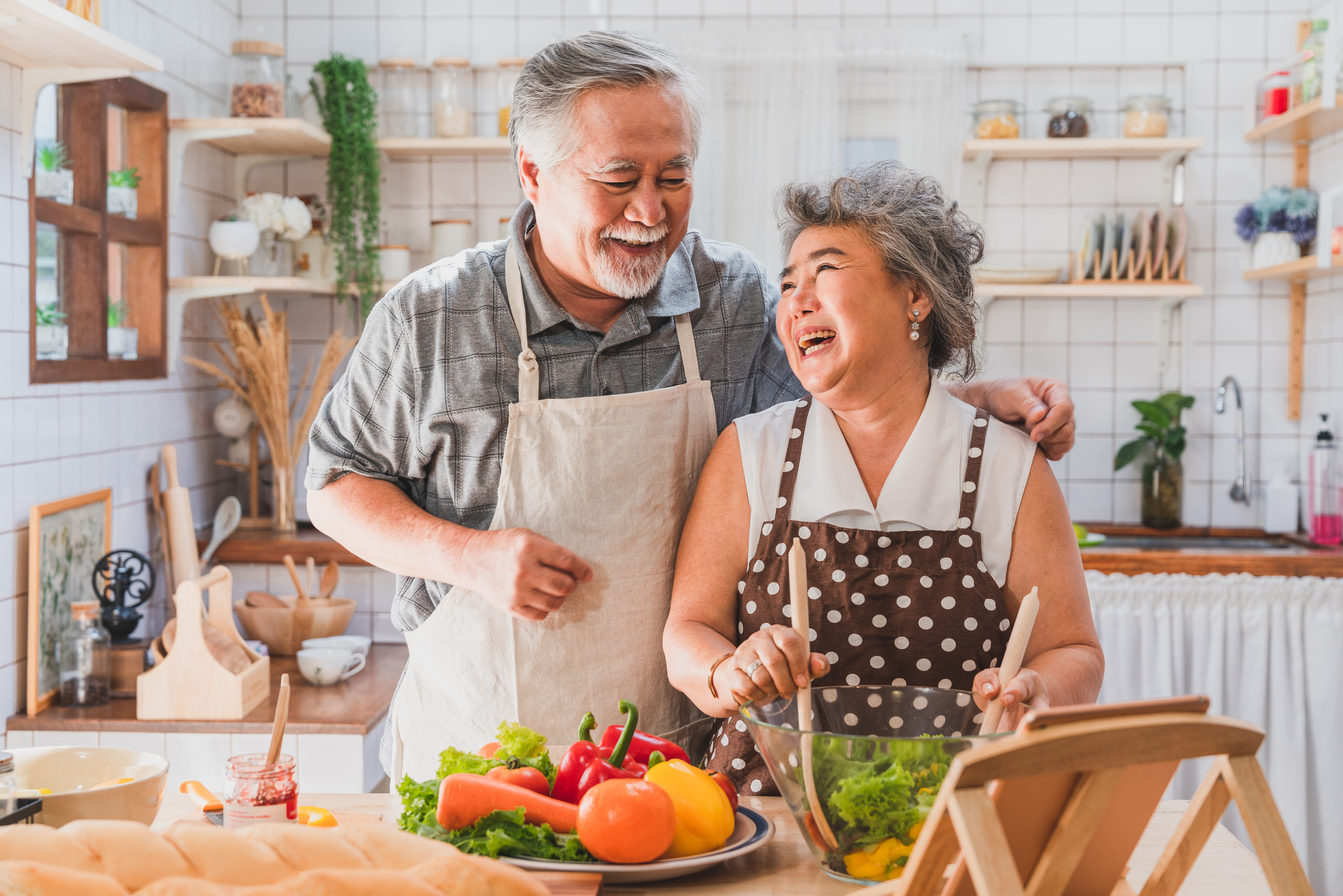  Healthy elderly couple smiling at each other and making a salad in the kitchen.