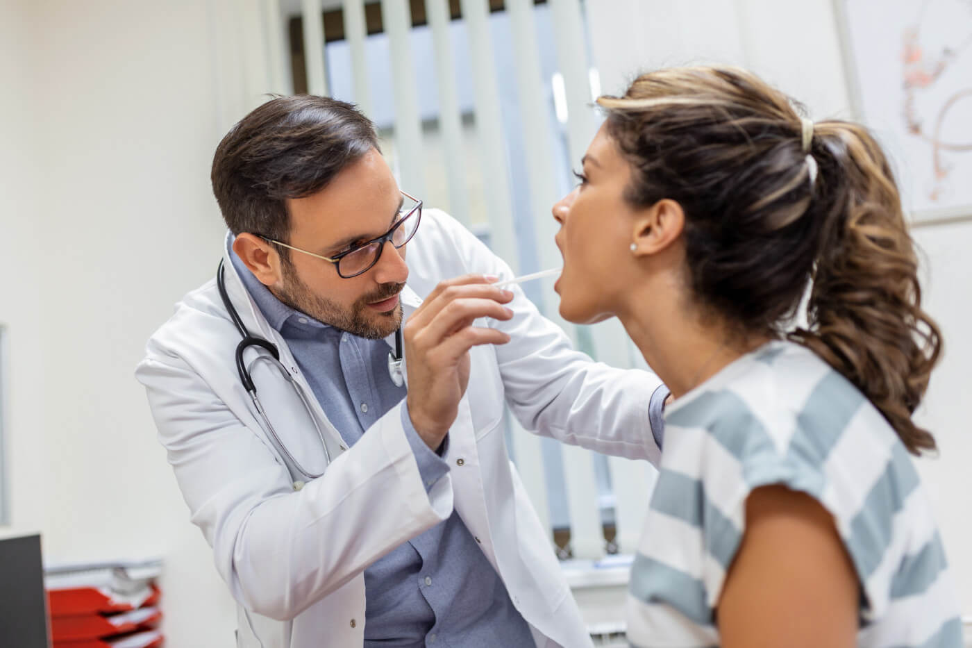 Doctor performing an oral exam on a female patient, using a tongue depressor to inspect her tongue. 